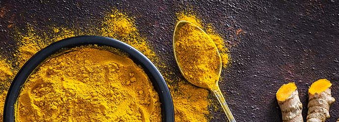 Vista de cima de uma tigela preta cheia de cúrcuma em pó em cima de mesa rústica. Uma colher de metal com pó de cúrcuma está ao lado da tigela e espalhado sobre a mesa. Raízes de cúrcuma fresca estão ao lado da colher.