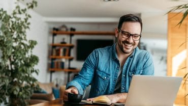 homem sorri em home office, olhando para laptop e bebendo xícara de café.