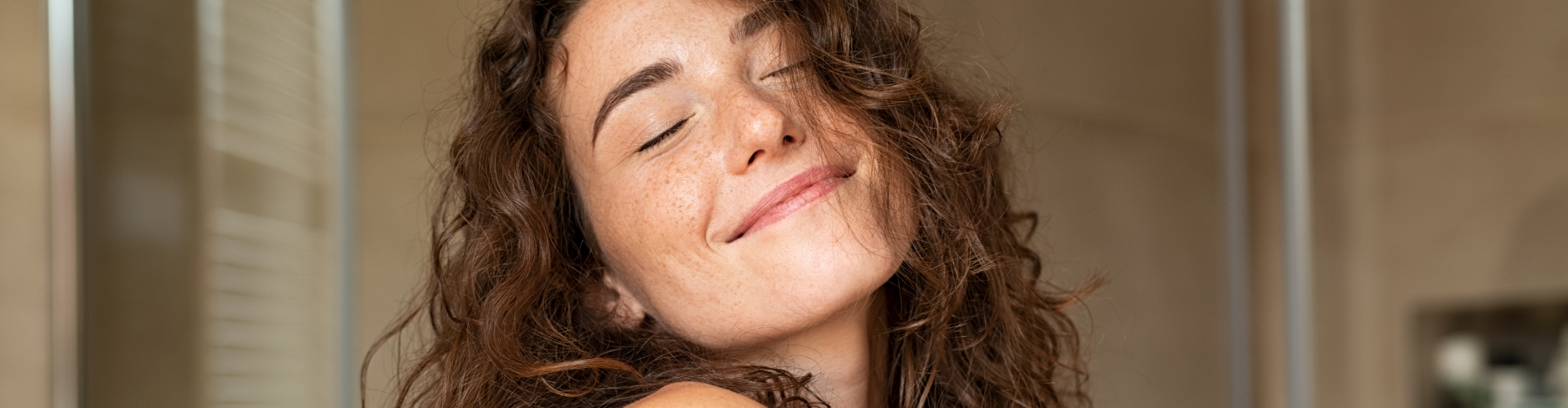 Mulher com os cabelos ondulados está de olhos fechados e sorrindo serenamente enquanto se abraça.