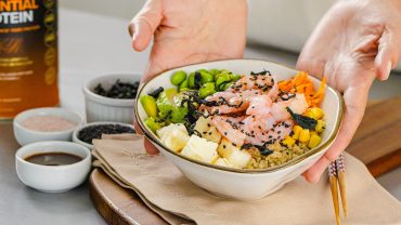 prato de poke havaiano de camarão, servido em uma tigela. O poke é composto por camarões, cubos de tofu, edamame, abacate, cenoura ralada, milho, algas marinhas e sementes de gergelim preto, dispostos sobre uma base de quinoa. Ao fundo, há potes com ingredientes e um suplemento proteico que complementa a composição visual da cena. Ideal para representar uma refeição saudável e nutritiva.